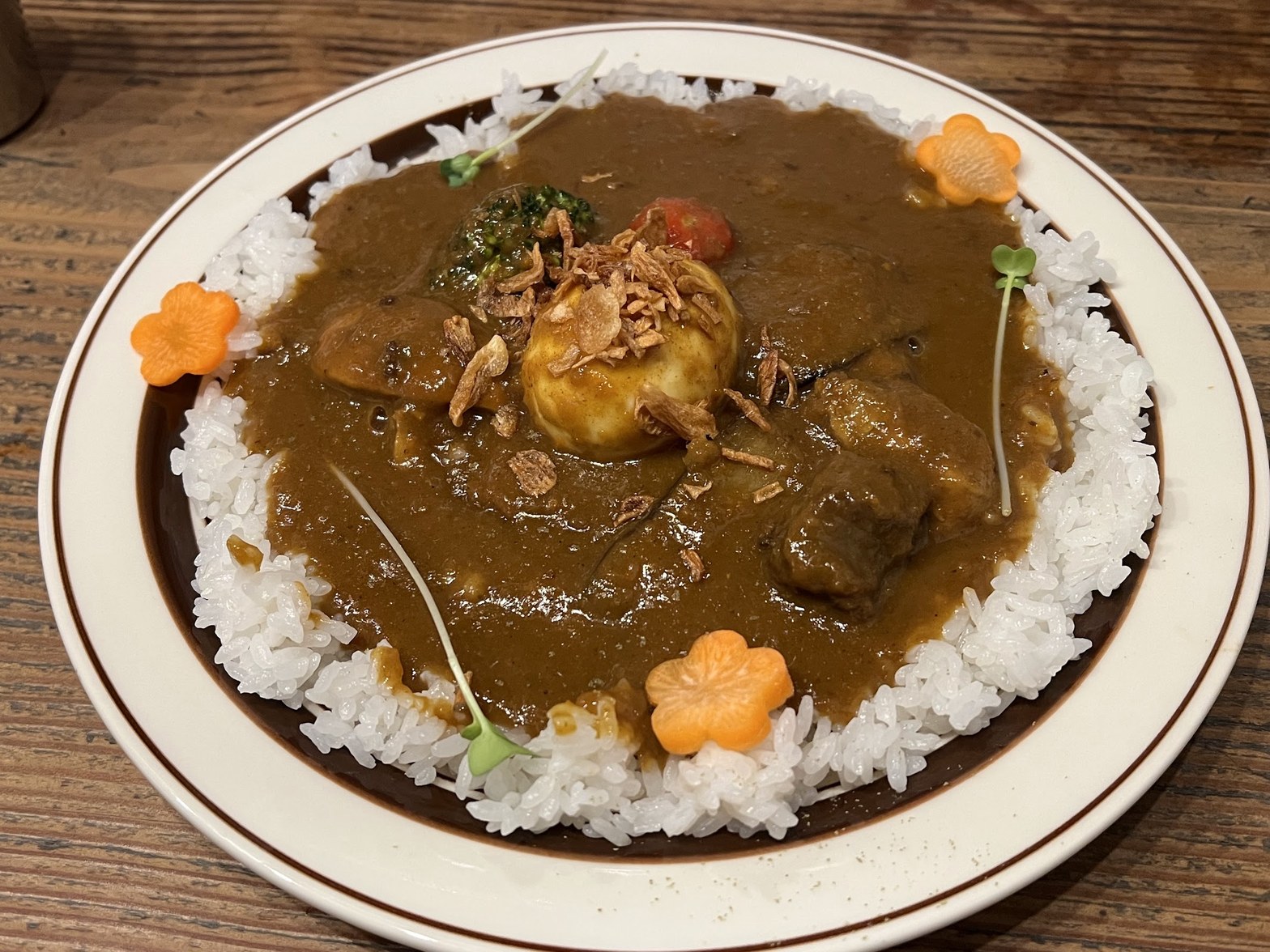 白いご飯を縁に盛りつけた丸皿に、チキン・ビーフ・やさい・ゆで卵を載せた濃い茶色の欧風カレーソース、上にフライドオニオンと花形にんじんが添えられている